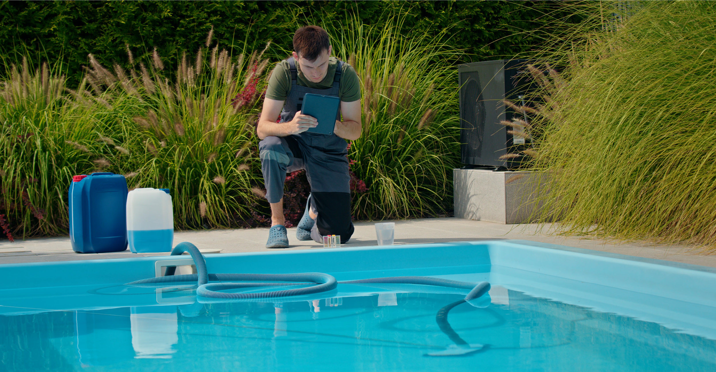 Professionnel de l'entretien de piscine vérifiant des données sur une tablette près d'une piscine coque, entouré de végétation luxuriante, avec accessoires de nettoyage et bouteilles de produits chimiques visibles