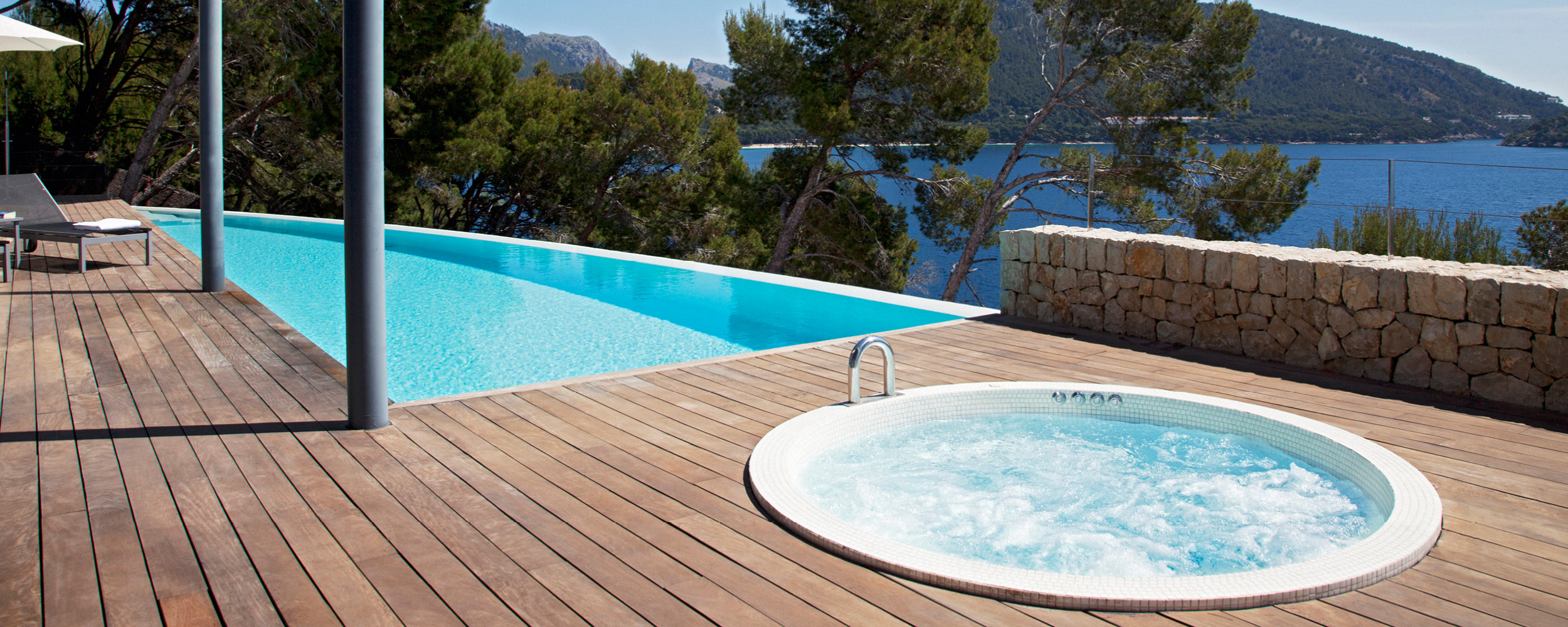 Piscine coque à débordement bleu turquoise avec spa, entourée d'une terrasse en bois exotique et chaises longues, offrant une vue sur la nature et un paysage côtier verdoyant.