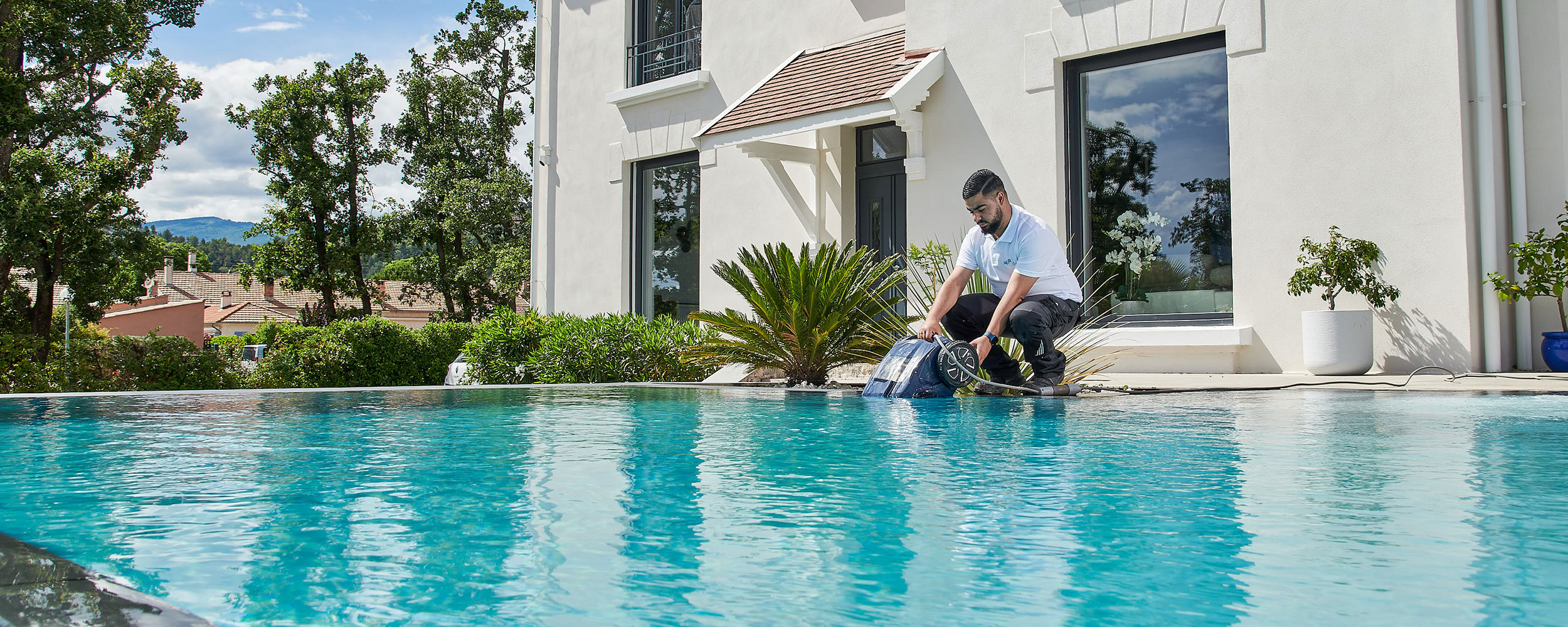 Professionnel en entretien de piscine travaillant sur une piscine coque moderne avec eau turquoise, située dans un quartier résidentiel avec maisons modernes en arrière-plan, plantes décoratives et debordement élégant, un environnement extérieur idyllique par une journée ensoleillée.