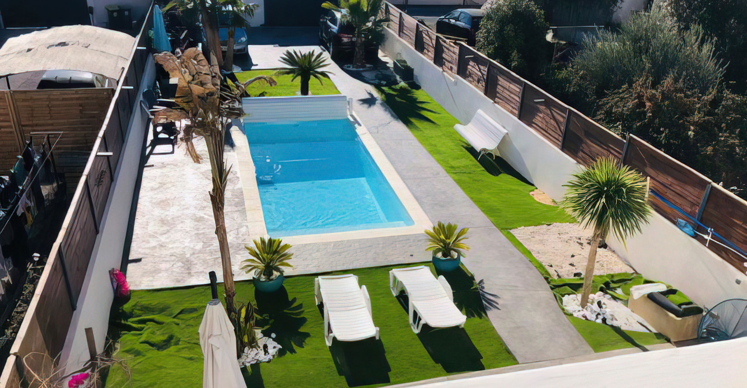 Espace extérieur aménagé avec piscine coque rectangulaire entourée de gazon synthétique, chaises longues blanches, et végétation tropicale, offrant un cadre idyllique et fonctionnel pour la détente.